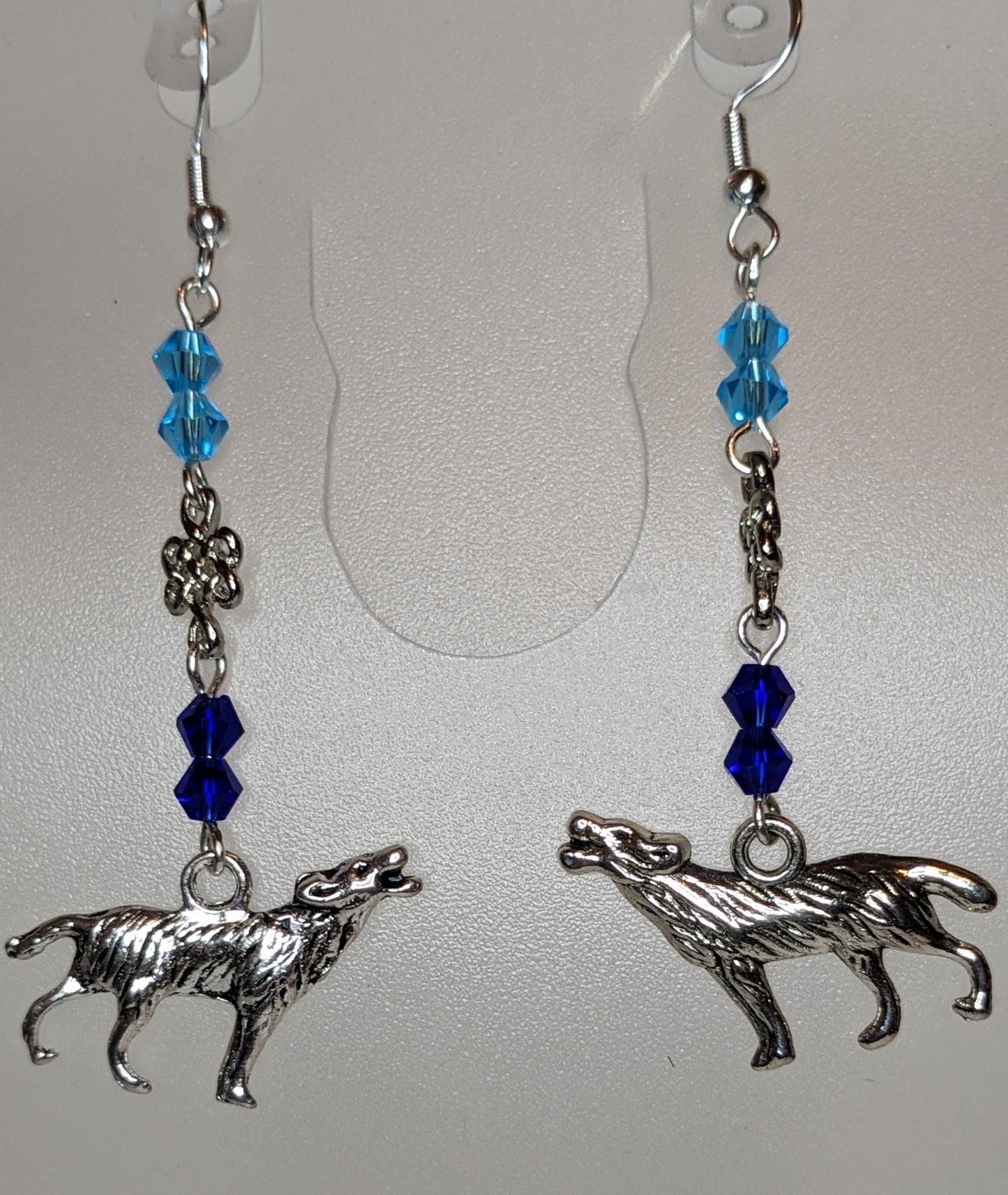 Close up of a pair of earrings with silver howling wolf charms at the bottom and faceted bicone crystal beads in light blue & dark blue separated by a silver Celtic knot connector. 