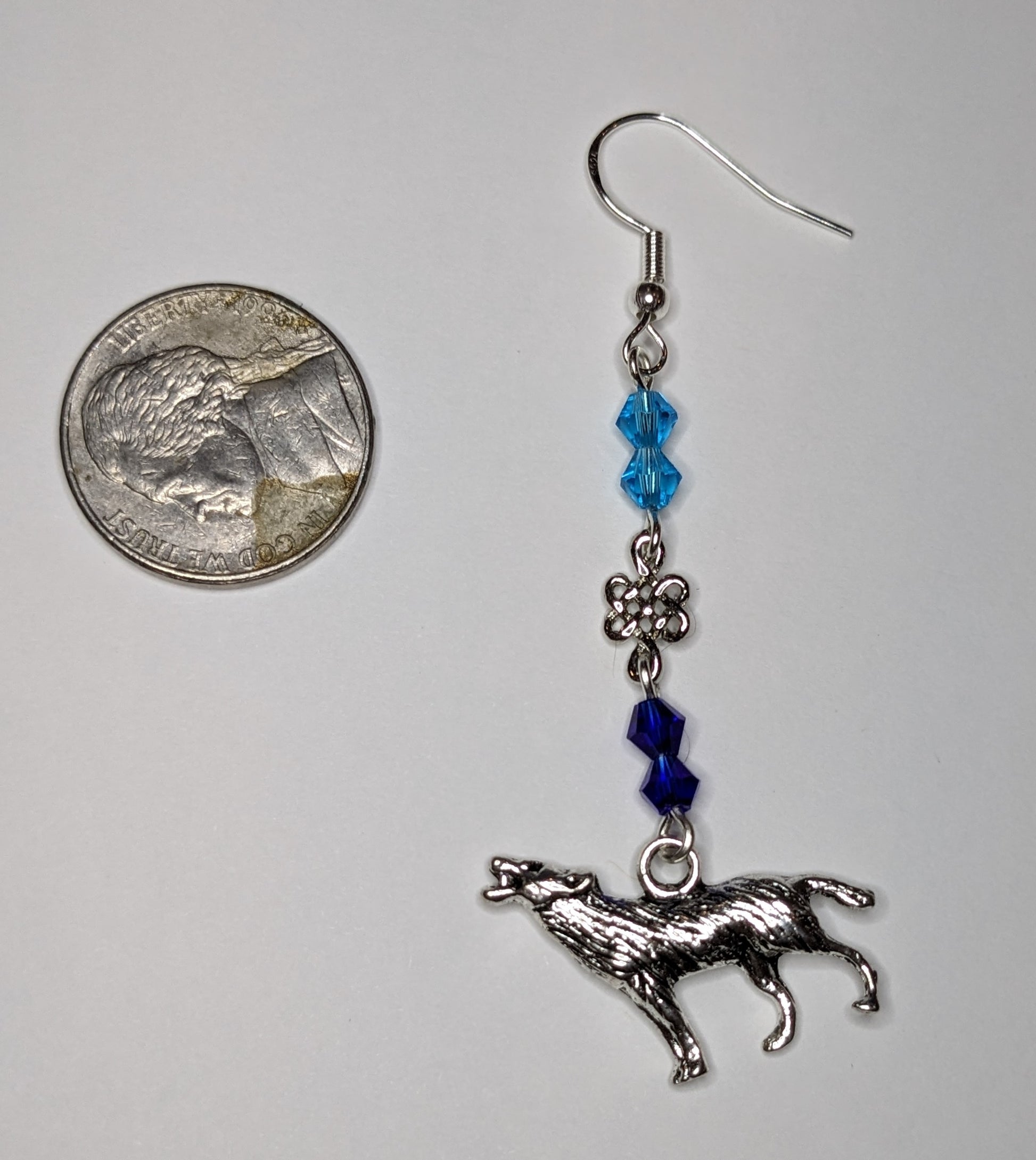 Close up of a single earring with silver howling wolf charm at the bottom and faceted bicone crystal beads in light blue & dark blue separated by a silver Celtic knot connector. Earring is next to a nickel to show scale 