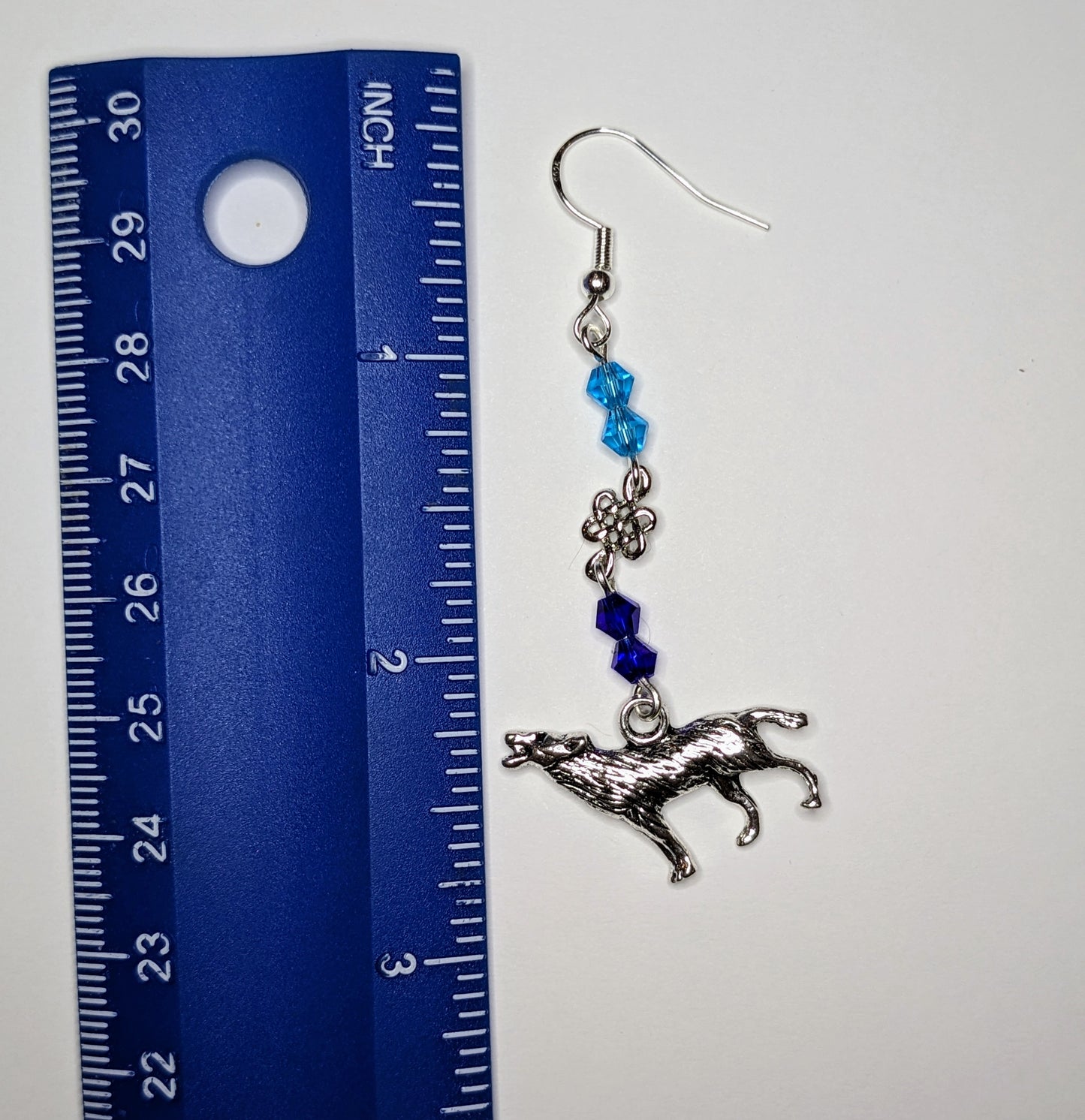 Close up of a single earring with silver howling wolf charm at the bottom and faceted bicone crystal beads in light blue & dark blue separated by a silver Celtic knot connector. Earring is next to a blue ruler and is 3 inches long 
