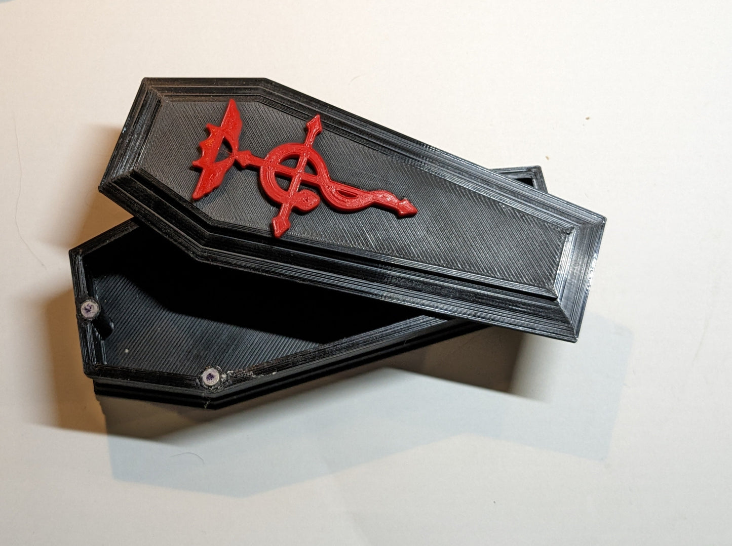 3D printed coffin shaped dice/trinket box in black with a red alchemy symbol in the top.  The top is slightly askew to show the inside of the box