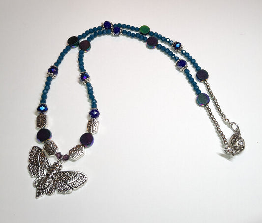 Beaded necklace with antiqued-silver colored deaths-head moth pendant.  The necklace has glass beads in shades of blue & teal with silver bead caps and engraved silver accent beads.  It has an ornate round silver magnetic clasp.  The necklace is about 20 inches long.