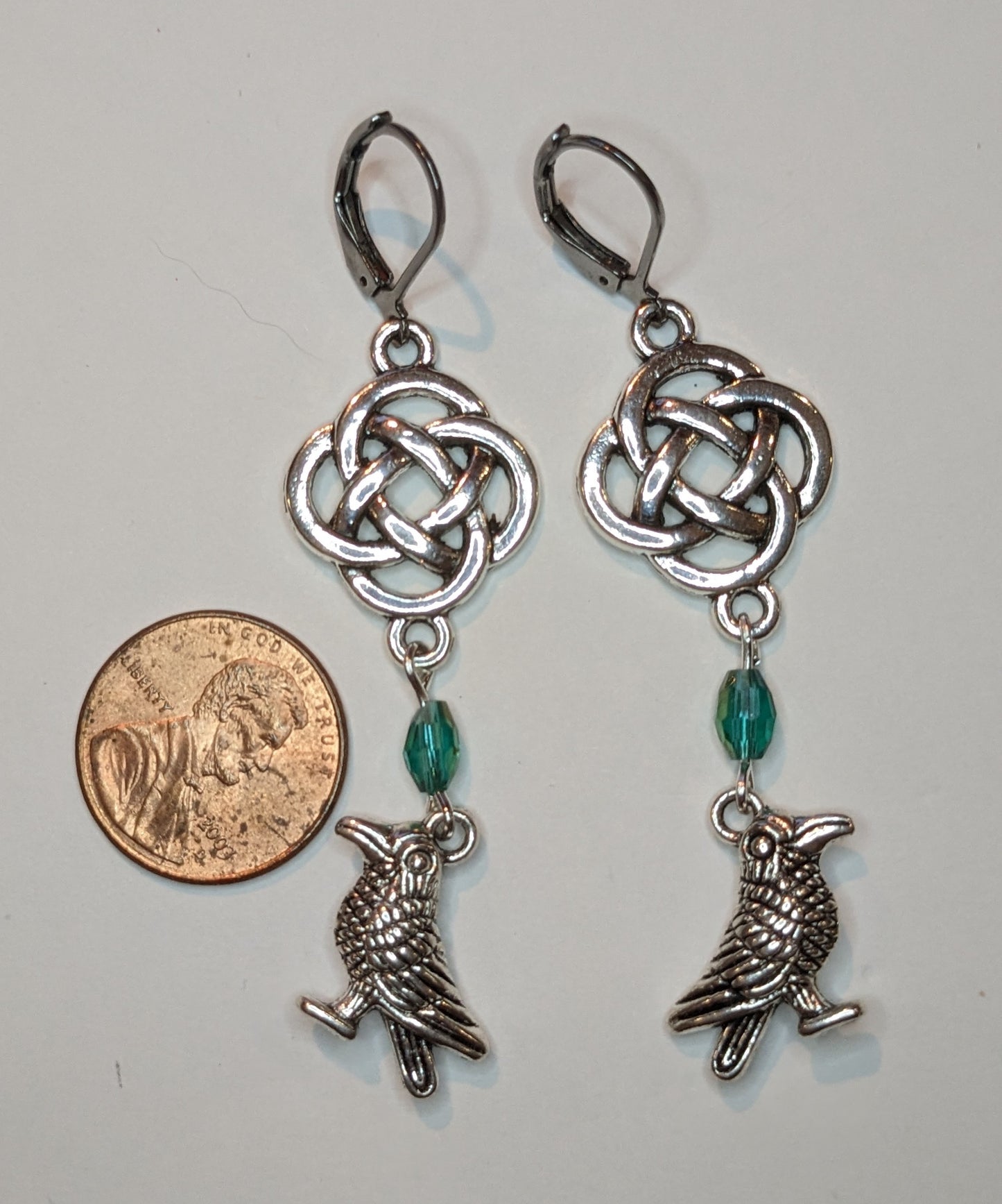 Earrings with silver toned raven charms, a blue - green crystal beads and a silver Celtic knot. A US penny sits to the left to show scale 