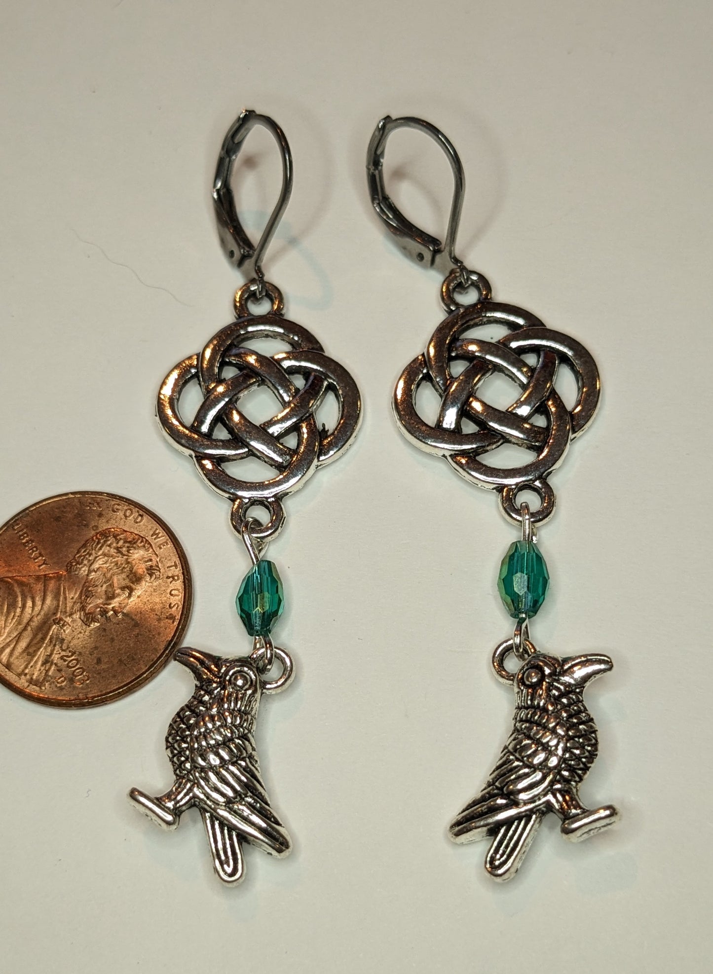 Earrings with silver toned raven charms, a blue - green crystal beads and a silver Celtic knot. A US penny is to the left to show scale 