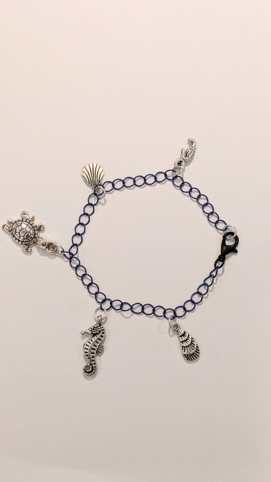 Charm bracelet with silver toned sea themed charms on a purple chain. There are five charms: 2 seahorses, 2 shells, and a turtle 