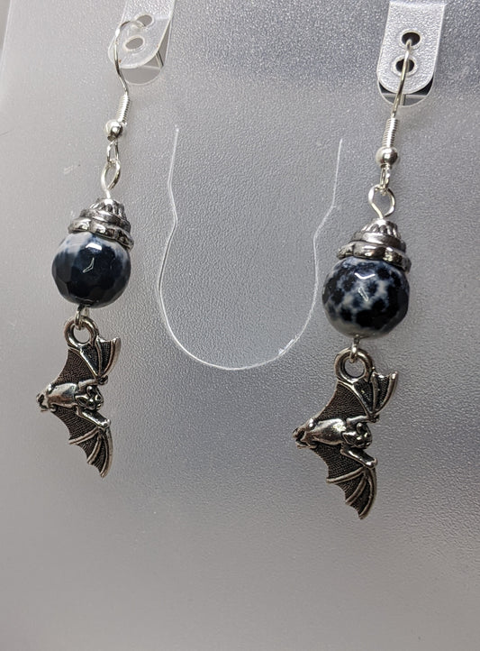 Pair of earrings on a white display.  Earrings are large faceted black and white beads with antiqued silver bead caps with cute silver bat charms at the bottom.  The bats are wings out. 
