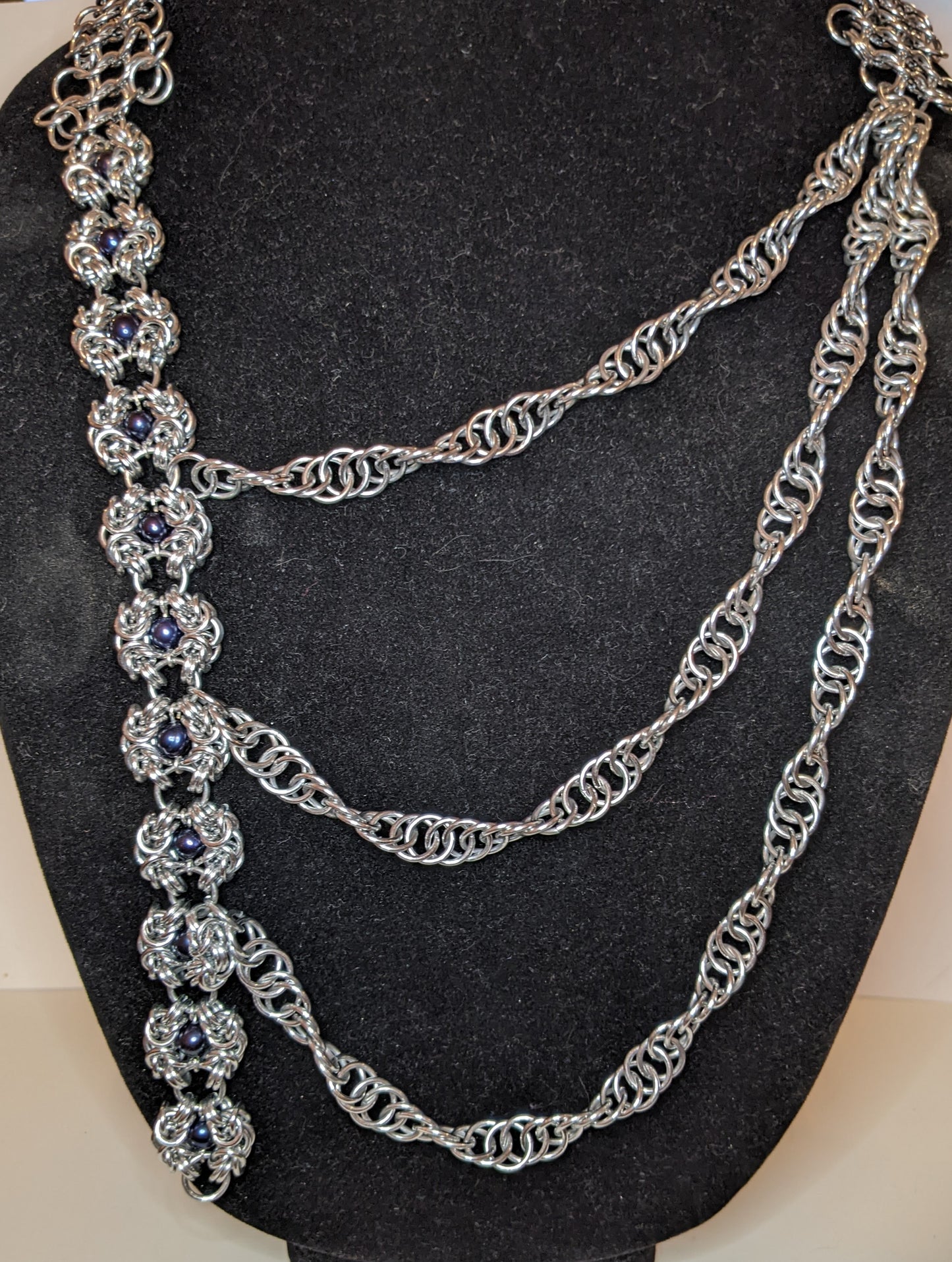 Silver asymmetrical chainmail necklace. At left are a series of Romanov weave flowers with black pearl bead centers, the right side is a series of three twisted ropes increasing in length that crosses over and attaches to the Romanov flowers. 