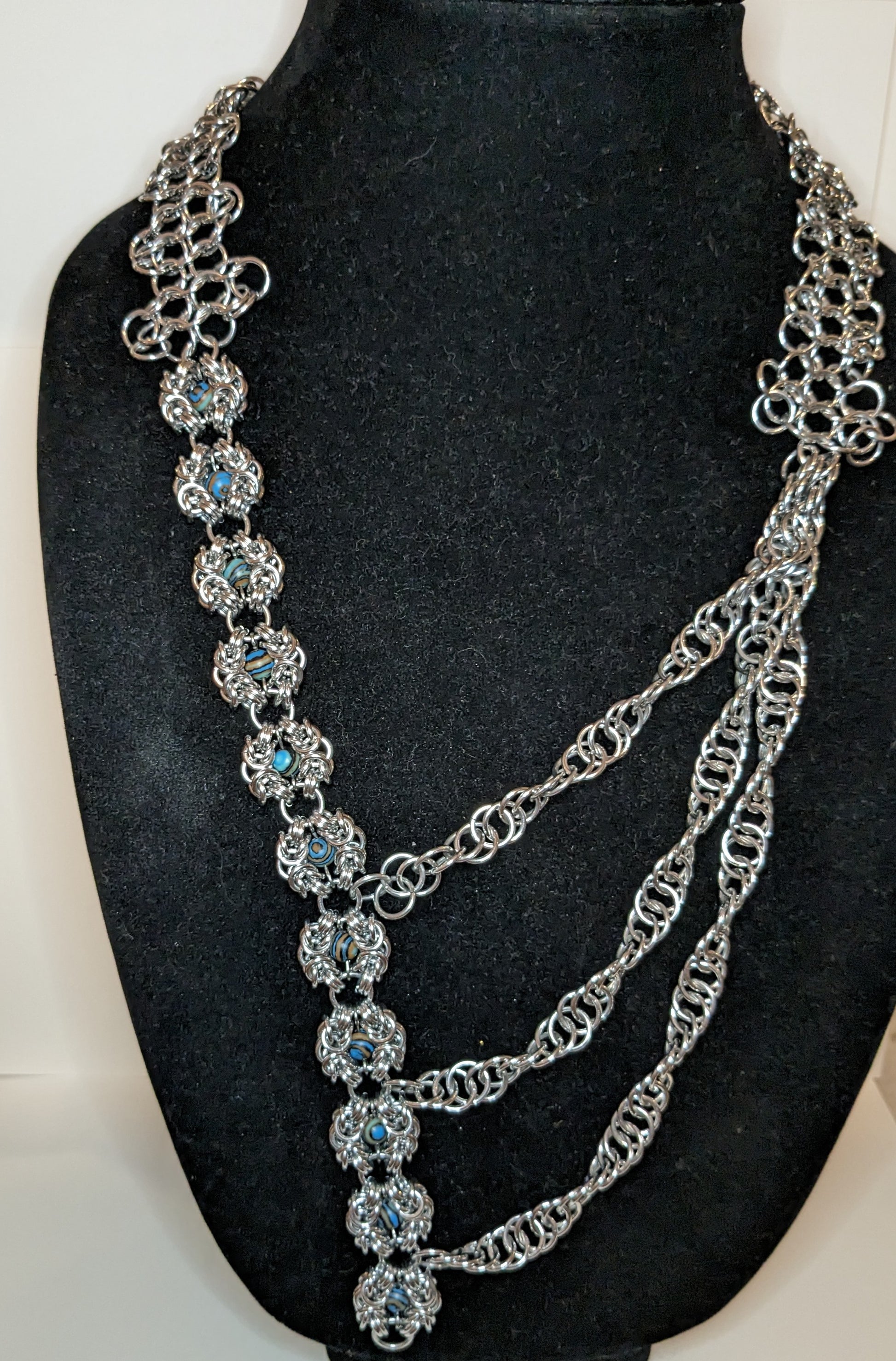 Asymmetric chainmail necklace on a black bust.  Left side is modified Romanov weave flowers with gemstone beads and the right side are three chainmail strands that cross the front to look like a mult-strand necklace.  