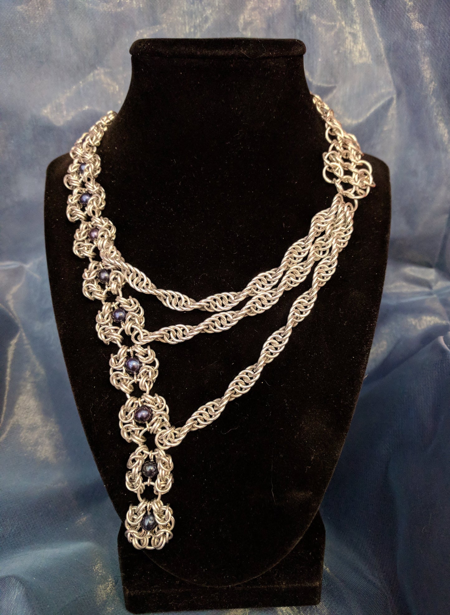 Silver asymmetrical chainmail necklace. At left are a series of Romanov weave flowers with black pearl bead centers, the right side is a series of three twisted ropes increasing in length that crosses over and attaches to the Romanov flowers. 
